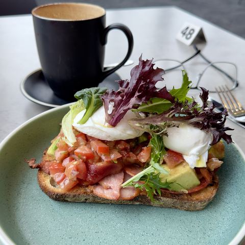 Breakfast toast topped with poached eggs, bacon, tomato and avocado, coffee alongside (mood photo)
