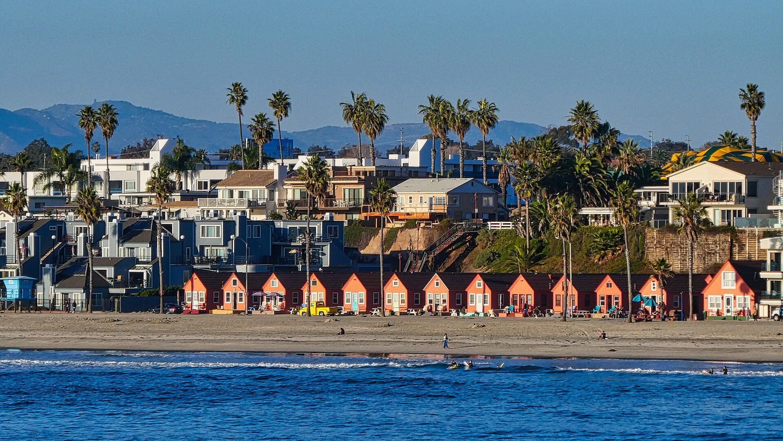 Oceanside beach seen from the pier: a row of orange cottages under palm trees with hills behind
